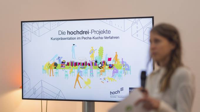 Dunkelblonde Frau mit Mikrofon neben einem Screen mit dem Text "Die hochdrei-Projekte. Kurzpräsentation im Pecha-Kucha-Verfahren" (öffnet Vergrößerung des Bildes)