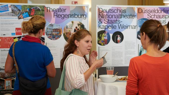 Zwei Personen unterhalten sich an einem Stehtisch, sie sind im Profil zu sehen. Eine dritte Person steht mit dem Rücken zur Kamera vor twei Flipchart-Wänden und schaut sich bunt gestaltete Poster an, auf denen Kulturinstitutionen wie das Theater Regensburg oder das Düsseldorfer Schauspielhaus ihre Projekte vorstellen. (öffnet Vergrößerung des Bildes)