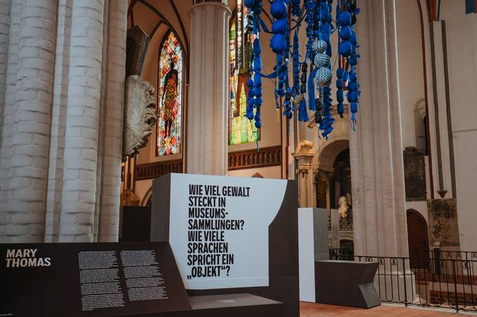 Blick in den Innenraum einer Kirche mit Säulen und einer Kunstinstallation, die von der Decke hängt sowie großer Texttafel auf der Folgendes zu Lesen ist: "Wie viel Gewalt steck in Museumssammlungen? Wie viele Sprachen spricht ein Objekt?"