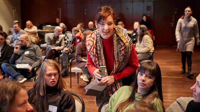 Im Vordergrund sitzt eine Gruppe in einem Stuhlkreis und diskutiert, hinter ihnen beugt sich eine stehende Person und spricht zu ihnen. (öffnet Vergrößerung des Bildes)