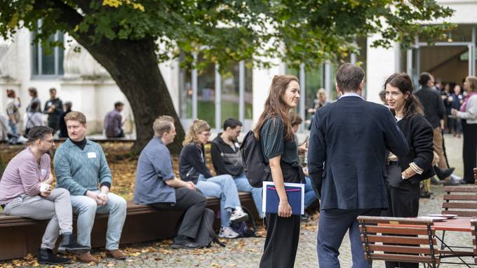 Menschen sitzen und stehen draußen unter einem großen Baum (öffnet Vergrößerung des Bildes)