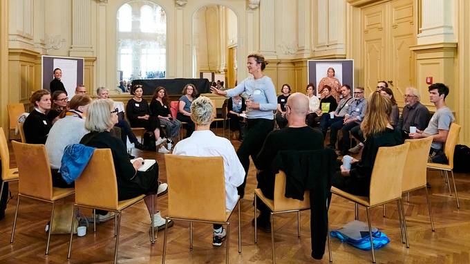 Etwa 40 Personen sitzen in einem Stuhlkreis in einem barocken Saal, in der Mitte steht eine Person und adressiert die Zuhörenden. Im Hintergrund stehen jeweils zwei kleinere Gruppen vor Flipchart-Präsentationen. (öffnet Vergrößerung des Bildes)