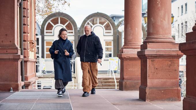 Zwei Personen gehen vor einem Gebäude mit Säulen entlang, sie tragen Winterkleidung und sind in ein Gespräch vertieft. (öffnet Vergrößerung des Bildes)