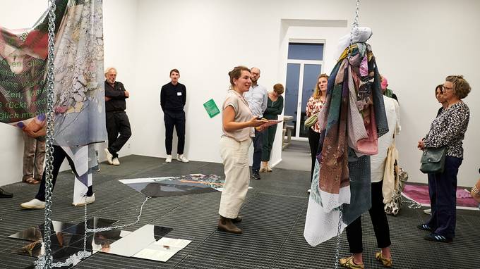 Eine Gruppe von Personen steht in einem Raum, der von einer größeren Installation ausgefüllt wird: An der Decke hängen Ketten, an denen bunte Textilien befestigt sind, auf dem Boden sind kachelartige Spiegel verteilt. Die Person in der Mitte erklärt der Gruppe die Hintergründe des Kunstwerks.. (öffnet Vergrößerung des Bildes)