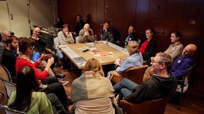 Eine Gruppe von Menschen sitzt im Kreis an einem Tisch und ist ins Gespräch vertieft. (öffnet Vergrößerung des Bildes)