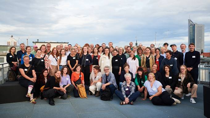 Ca. 80 Personen posieren für ein Gruppenbild auf einer Dachterrasse, im Hintergrund sind ein bewölkter Himmel und der MDR-Büroturm zu sehen. (öffnet Vergrößerung des Bildes)