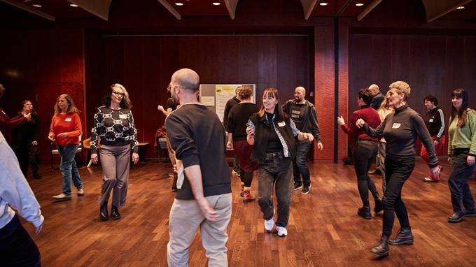 In einem Saal mit Parkettboden bewegt sich eine Gruppe von Menschen tanzend durch den Raum. (öffnet Vergrößerung des Bildes)